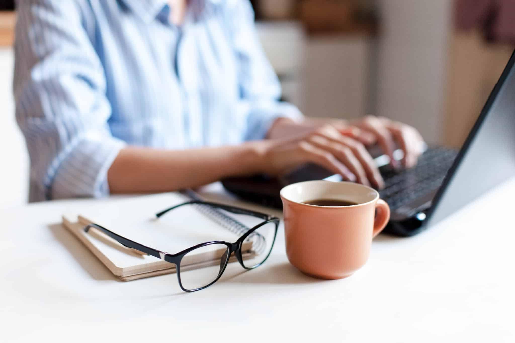 Remote working from home. Freelancer workplace in kitchen with laptop, cup of coffee, spectacles. Concept of distance learning, isolation, female business, shopping online. Close up of woman hands.