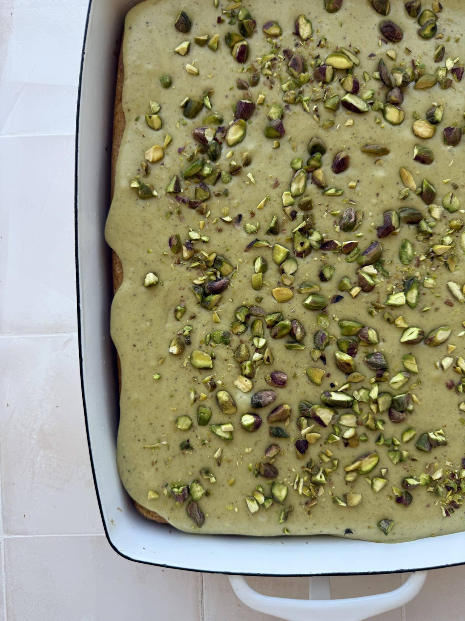 Ein frisch gebackener Pistazienkuchen mit cremiger, hellgrüner Glasur, bestreut mit gehackten Pistazien. Ein Stück ist bereits herausgeschnitten und zeigt die saftige Textur.