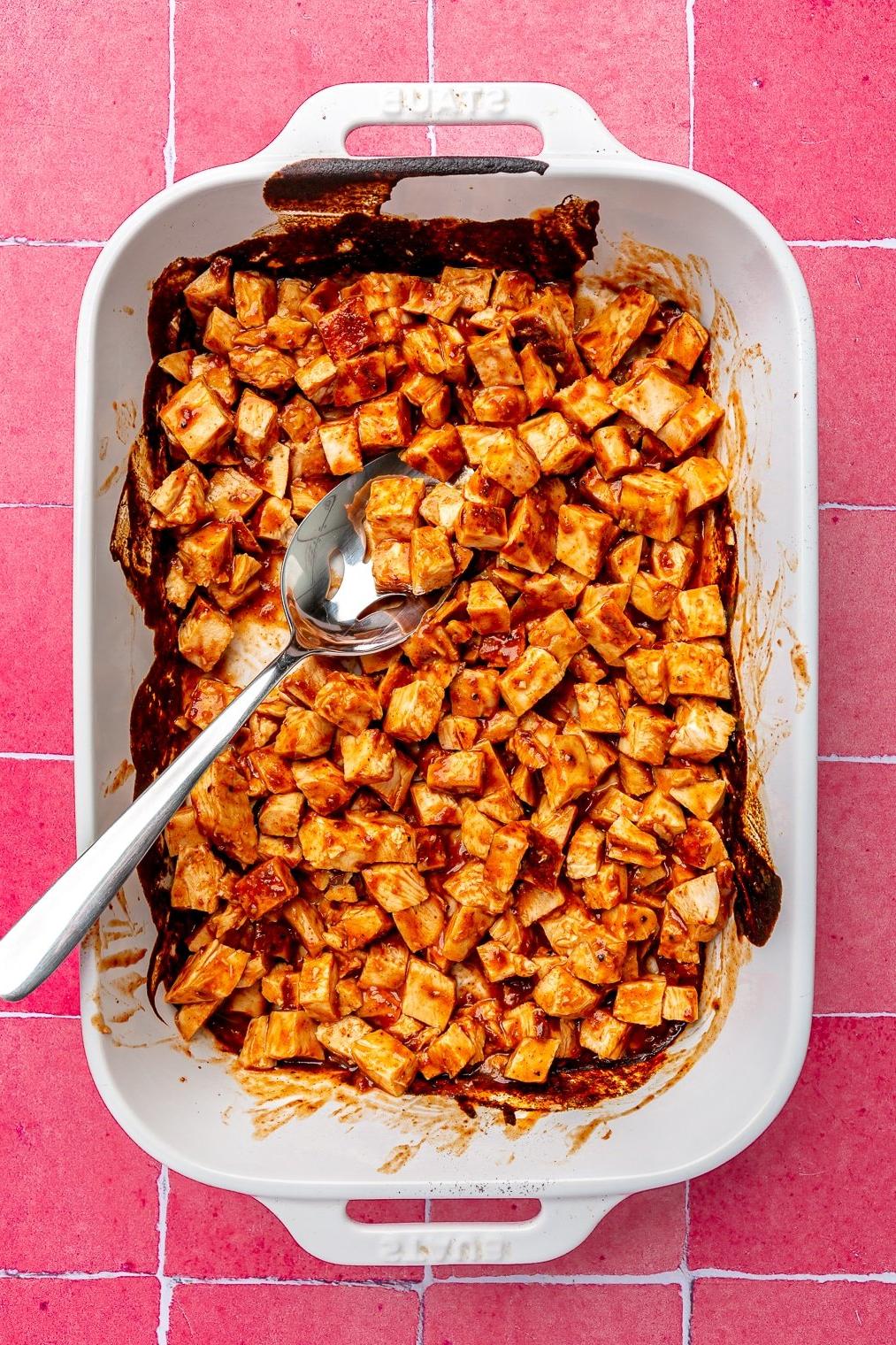 Eine weiße Schale mit gewürfelten BBQ-Hähnchenbrüsten auf einem pinkfarbenen Fliesenuntergrund.