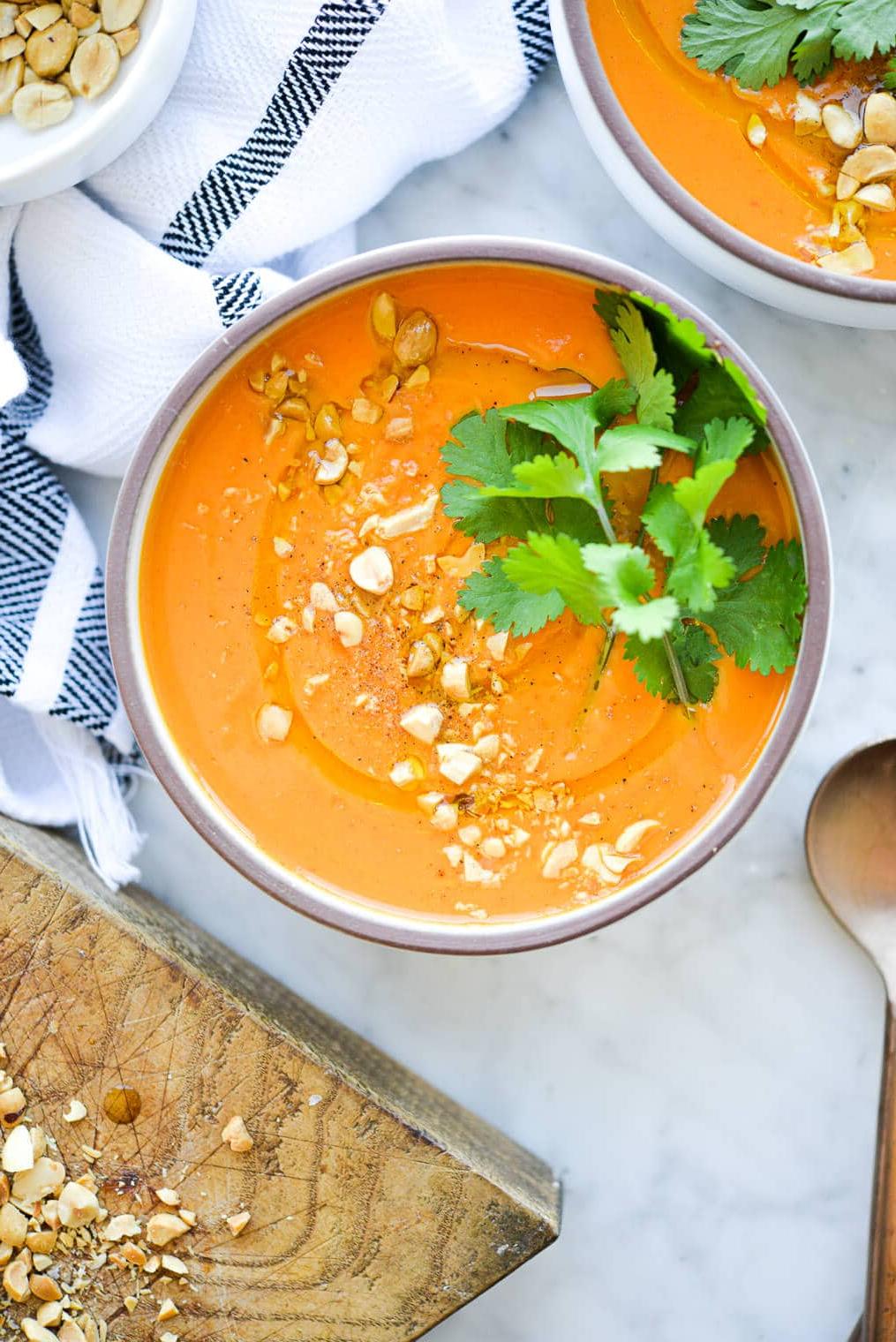 Eine Schüssel mit leuchtend oranger Thai-Karottensuppe, garniert mit frischem Koriander und zerdrückten Erdnüssen, auf einer Marmoroberfläche.