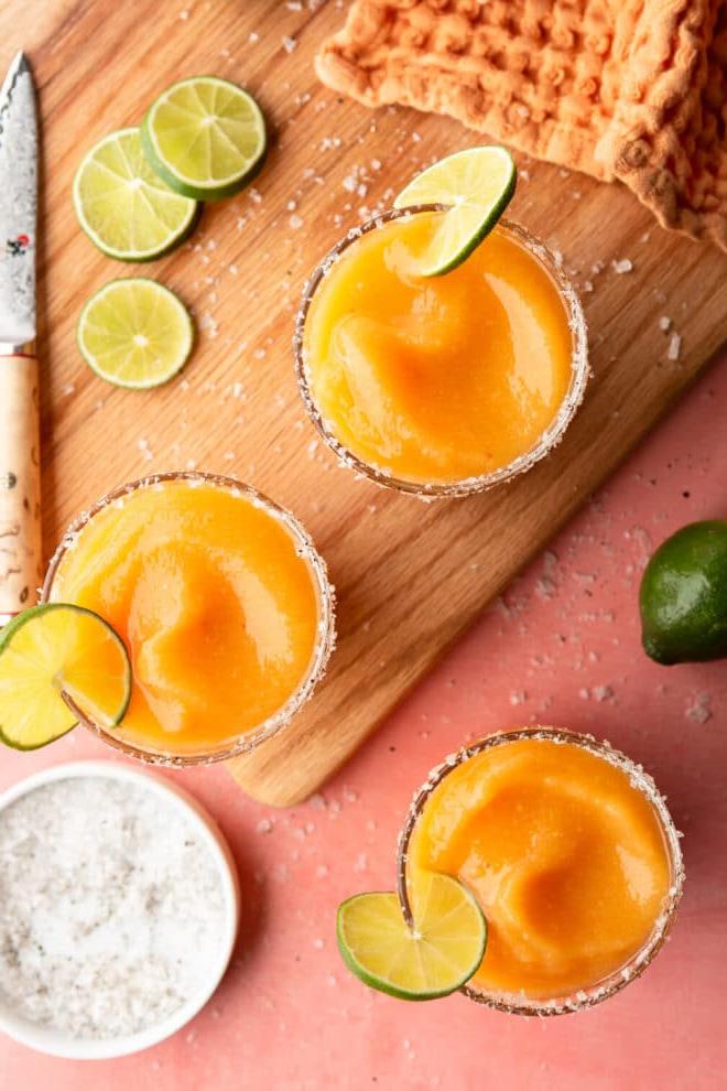 Aufnahme von oben: Gefrorene Pfirsich Margaritas in Gläsern, garniert mit Meersalzflocken und einer Limettenscheibe, auf einer pinkfarbenen Oberfläche mit Schneidebrett, Schälmesser, einer Prise Meersalzflocken, Limettenscheiben und orangefarbenem Leinentuch.