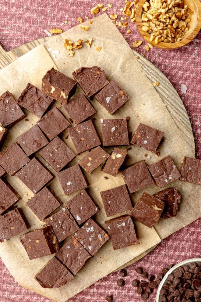 Hausgemachtes Schokoladen-Walnuss-Fudge 2 Schokoladen-Walnuss-Fudge in kleinen Quadraten auf braunem Backpapier auf einer dunkelrosafarbenen Tischdecke, daneben eine Holzschale mit Walnüssen und eine kleine Schüssel mit Schokoladenstückchen.