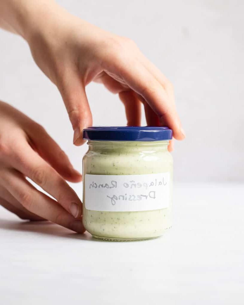 Eine Person hält ein kleines, beschriftetes Glas mit selbstgemachtem Jalapeño Ranch Dressing in den Händen.