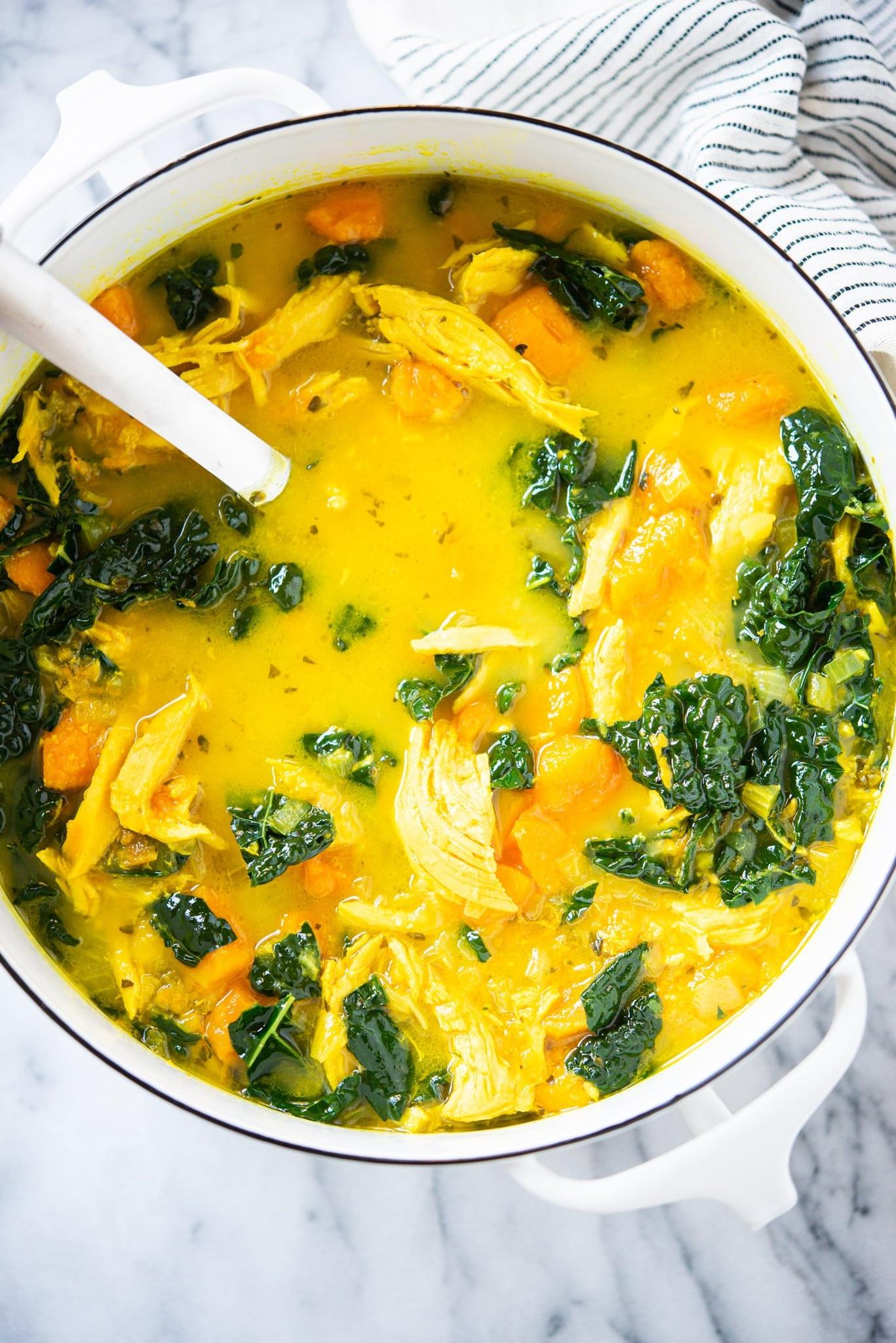 Gelbe Kurkuma-Hühnersuppe mit Süßkartoffeln und Grünkohl in einem weißen Topf auf einer Marmoroberfläche