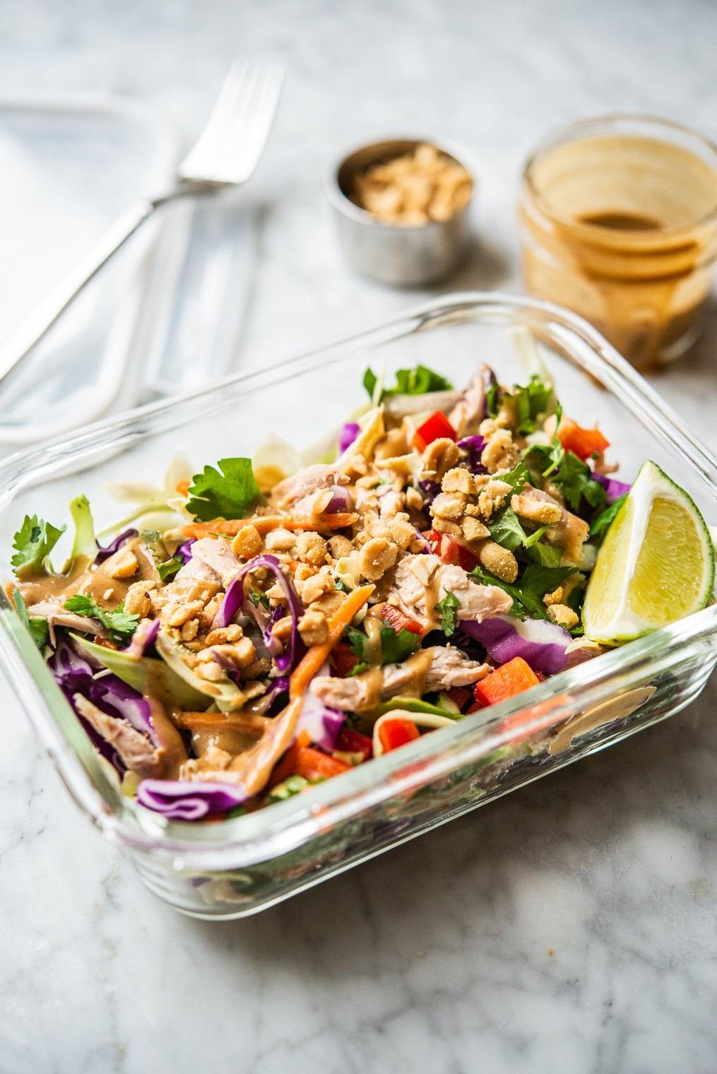 Ein farbenfroher Thai-Hähnchensalat in einem einzelnen Glas-Meal-Prep-Behälter mit Erdnussdressing und zerstoßenen Erdnüssen im Hintergrund