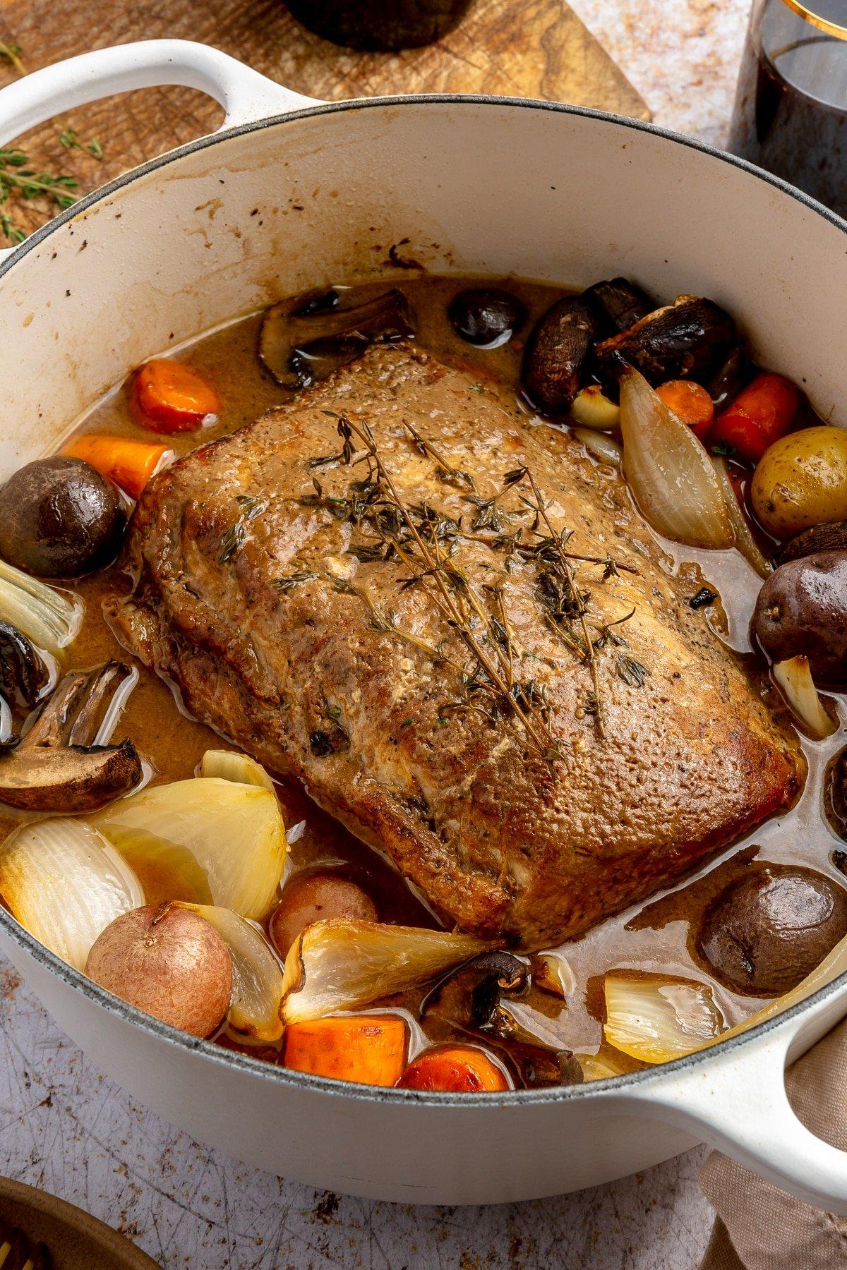 Ein fertig gegarter Balsamico-Schweinebraten liegt in einem weißen, emaillierten Topf. Zwei Weingläser, ein brauner Teller mit goldenem Besteck und ein Schneidebrett stehen daneben.