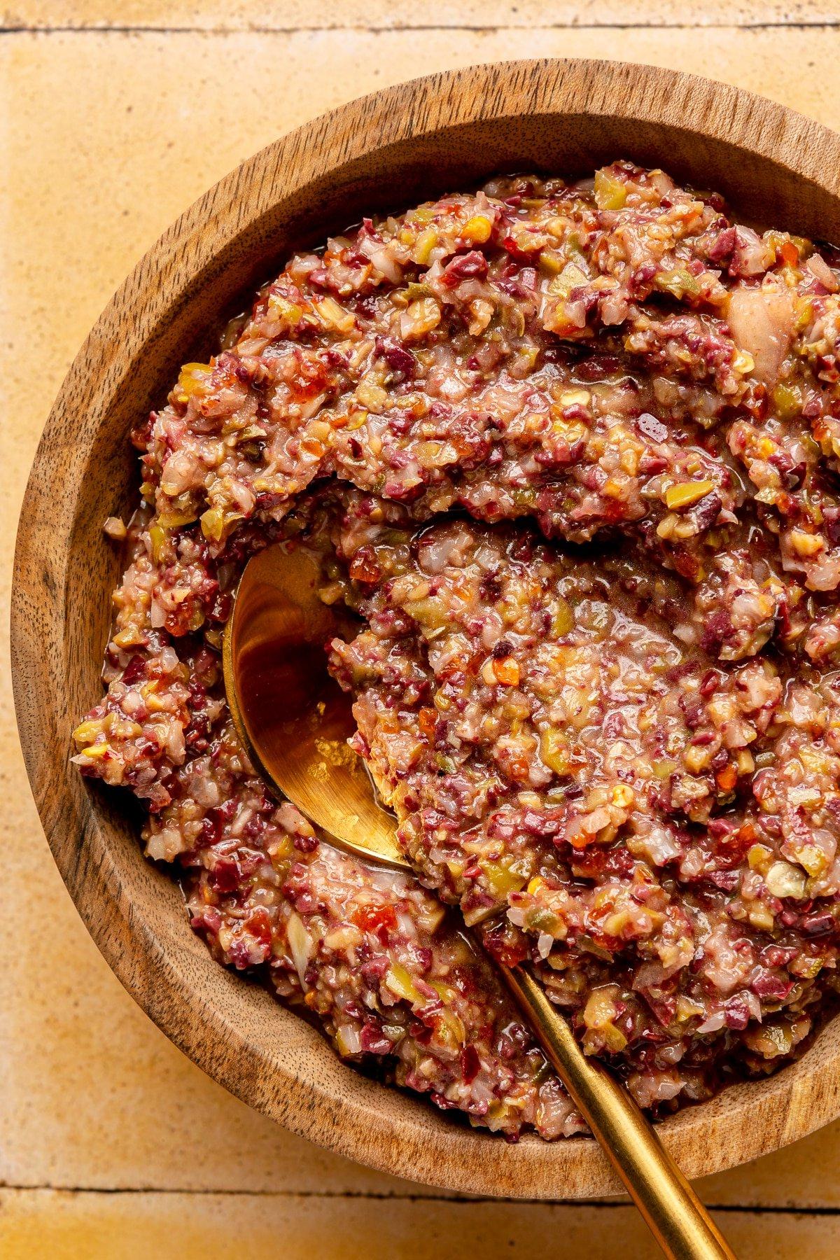Oliven-Tapenade in einer hölzernen Servierschale. Ein goldfarbener Löffel liegt daneben.