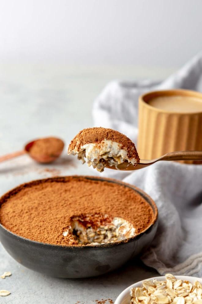 Eine seitliche Ansicht einer Schale Tiramisu Overnight Oats in einer schwarzen Schale. Eine Hand hält den Löffel mit einem Löffel Haferflocken über der Schale. Im Hintergrund ist eine Tasse Kaffee zu sehen.