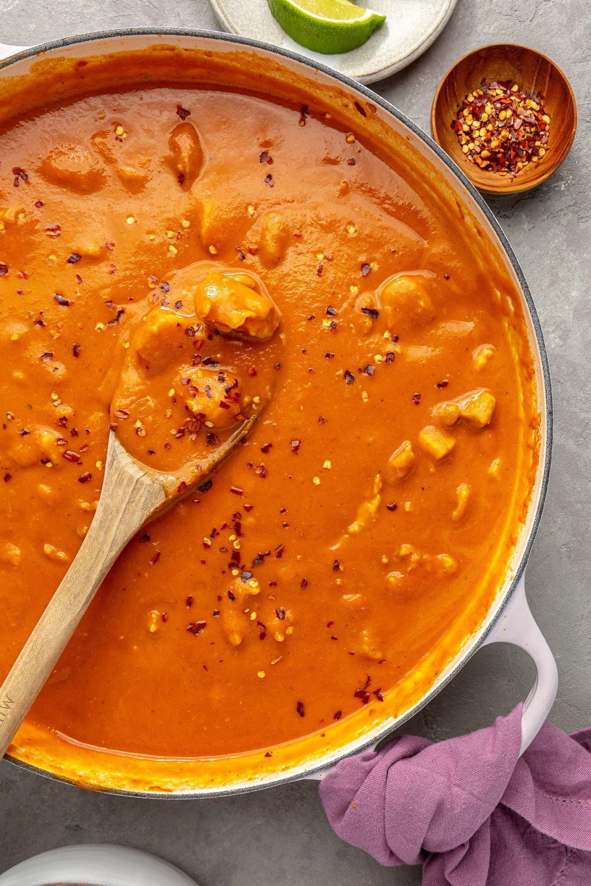 Einfaches Kürbiscurry-Rezept 2 Ein weißer Topf mit Kürbis-Curry, das mit roten Pfefferflocken bestreut ist. Ein Holzlöffel ist zu sehen, wie er einen Teil des Currys schöpft.