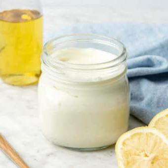 Glas mit hausgemachter Mayonnaise auf einer grau-weißen Theke mit Zitronenhälften, Avocadoöl im Hintergrund, einem Servierlöffel und einem hellblauen Leinentuch.