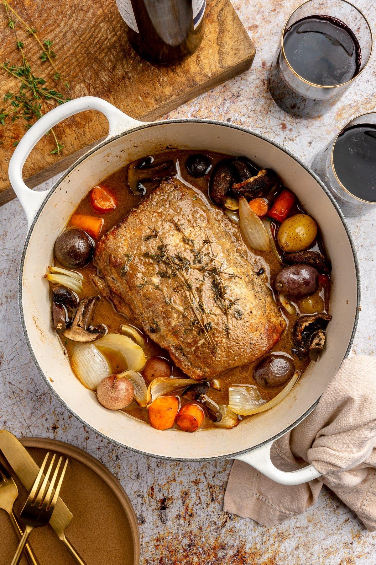 Ein fertig gegarter Balsamico-Schweinebraten liegt in einem weißen, emaillierten Topf. Zwei Weingläser, ein brauner Teller mit goldenem Besteck und ein Schneidebrett stehen daneben.