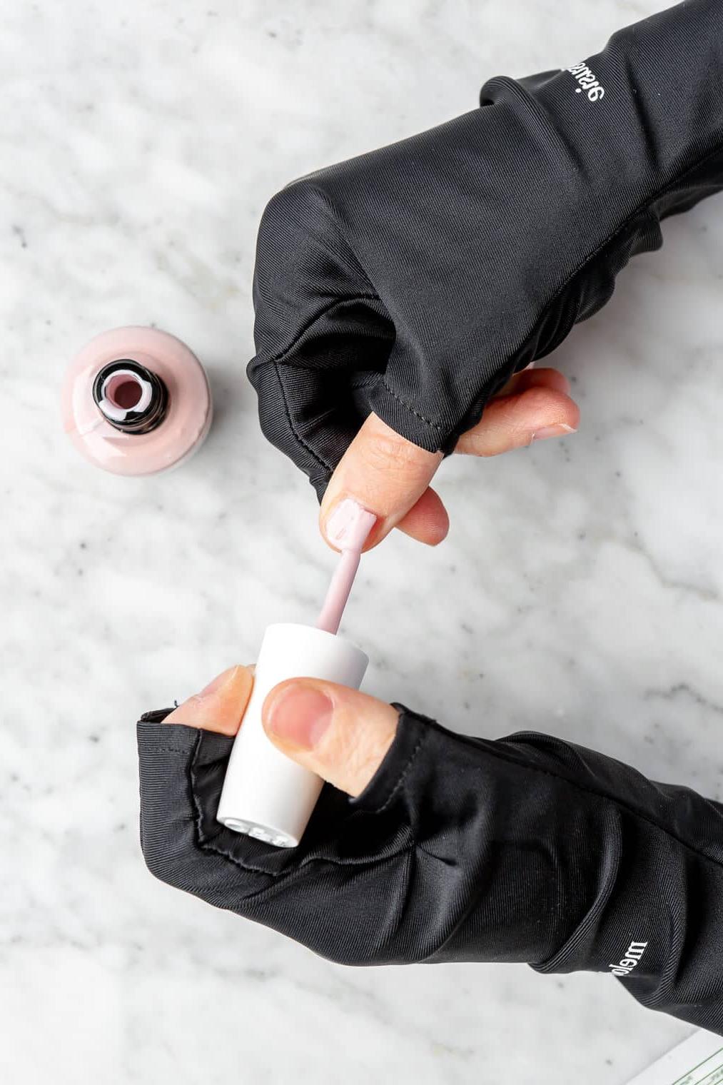 Hände tragen schwarze, fingerlose Handschuhe und tragen eine Schicht hellrosa Nagellack auf die Nägel auf.