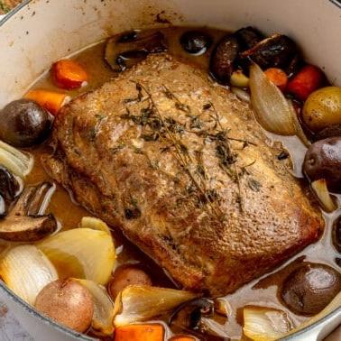 Ein fertig gegarter Balsamico-Schweinebraten liegt in einem weißen, emaillierten Topf. Zwei Weingläser, ein brauner Teller mit goldenem Besteck und ein Schneidebrett stehen daneben.