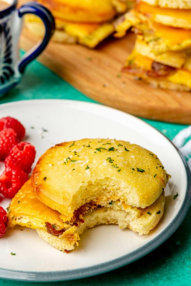 Ein Frühstücks-Sandwich auf einem Teller mit Himbeeren. Im Hintergrund befindet sich eine weiß-blaue Tasse und ein Holzschneidebrett mit weiteren Frühstücks-Sandwiches. Alles steht auf einer türkisfarbenen Oberfläche.