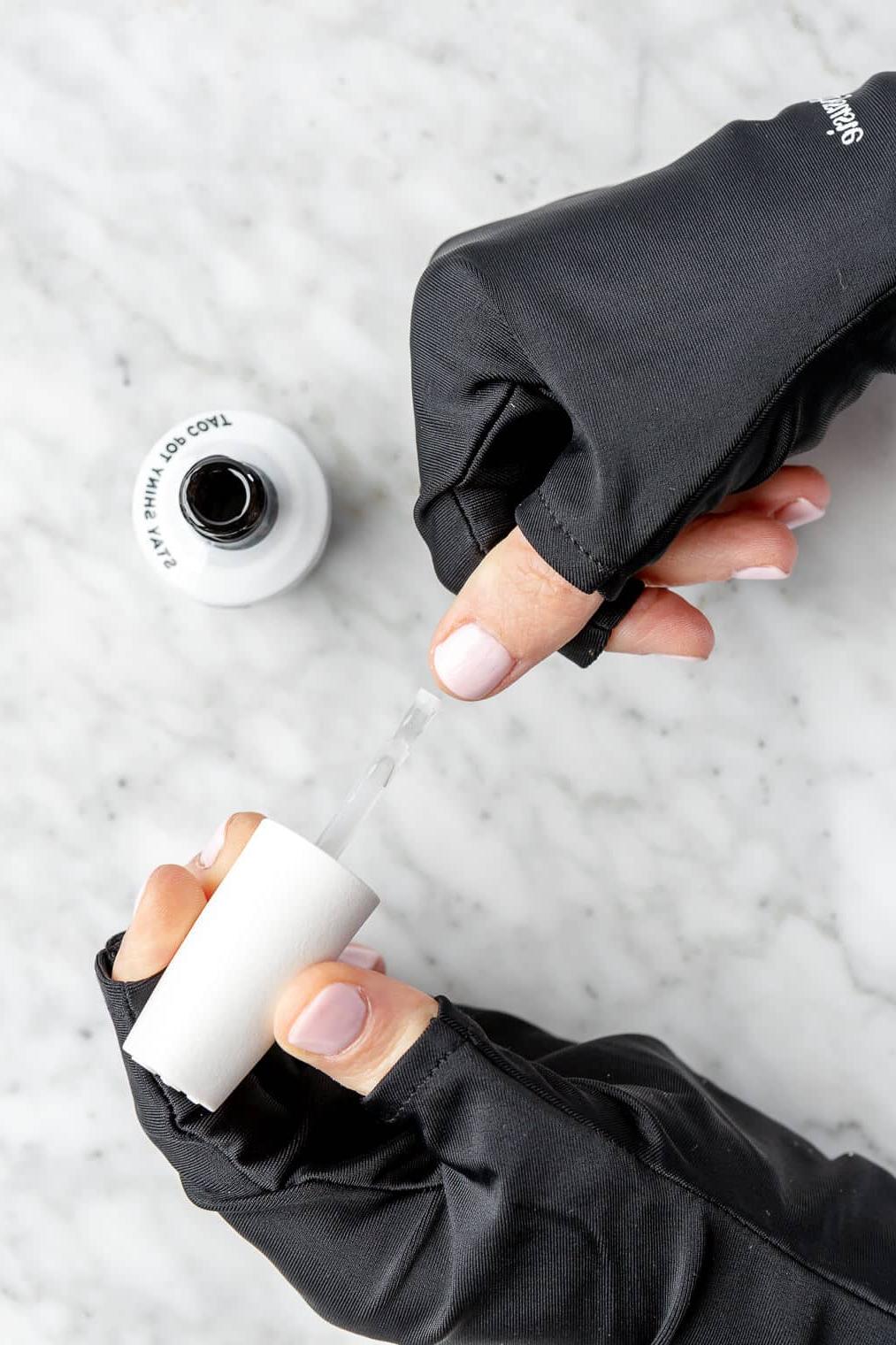 Hände tragen fingerlose schwarze Handschuhe und tragen eine Deckschicht Nagellack auf die lackierten Nägel auf.