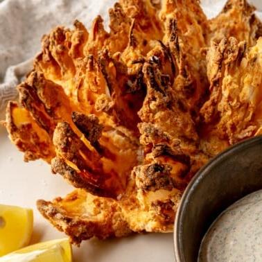 Eine blühende Zwiebel aus der Heißluftfritteuse liegt perfekt gegart und goldbraun auf einem Teller, daneben eine Schale mit cremiger Dip-Soße und frischen Zitronenspalten für eine appetitliche Präsentation.