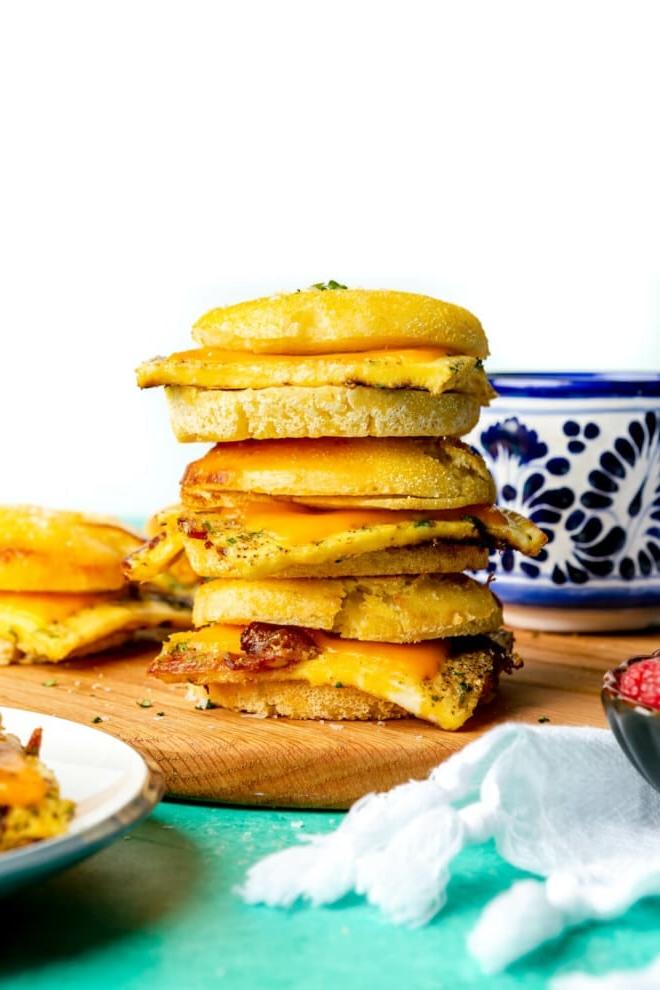 Stapel von 3 Frühstücks-Sandwiches auf einem Holzschneidebrett. Im Hintergrund befindet sich eine weiß-blaue Tasse.