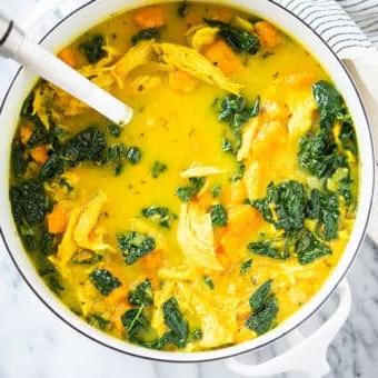 Gelbe Kurkuma-Hühnersuppe mit Süßkartoffeln und Grünkohl in einem weißen Topf auf einer Marmoroberfläche