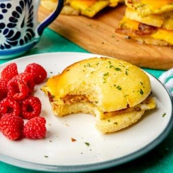 Ein Frühstücks-Sandwich auf einem Teller mit Himbeeren. Im Hintergrund befindet sich eine weiß-blaue Tasse und ein Holzschneidebrett mit weiteren Frühstücks-Sandwiches. Alles steht auf einer türkisfarbenen Oberfläche.
