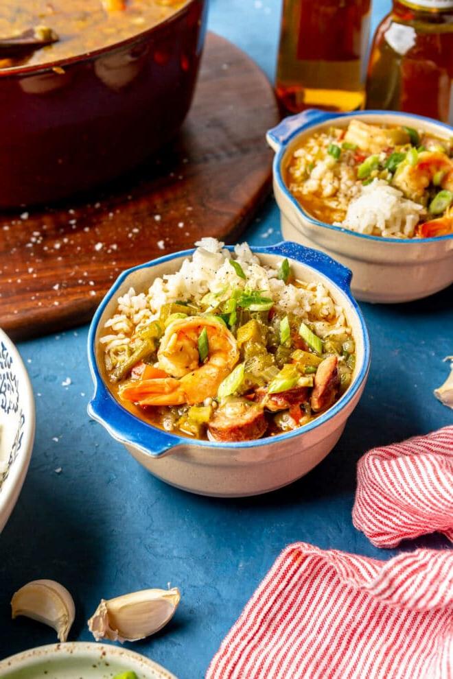Zwei blau umrandete Ramekins mit einer Portion weißem Reis und Garnelen- und Wurst-Gumbo. Sie stehen auf einem blauen Tisch mit einem Topf Gumbo im Hintergrund und einer rot-weiß dünn gestreiften Leinentischdecke davor.