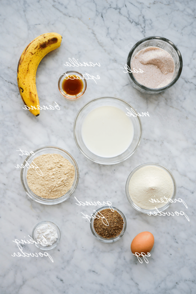 Beschriftete Zutaten für Protein-Pancakes in Schüsseln auf einer Marmoroberfläche.