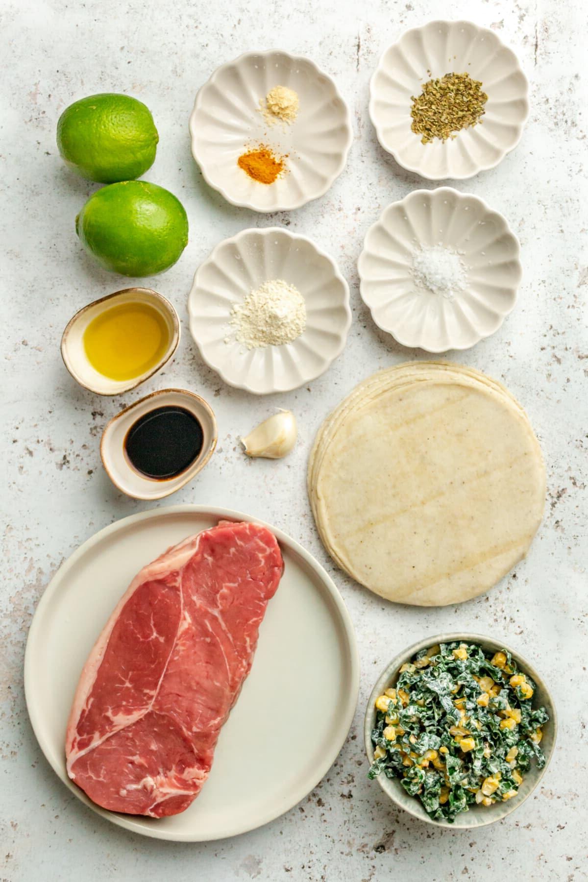 Zutaten für Carne Asada Tacos mit Adobo-Limetten-Marinade liegen auf verschiedenen kleinen Tellern und Schalen neben einem Grünkohl-Mais-Krautsalat auf einer hellgrauen Oberfläche.