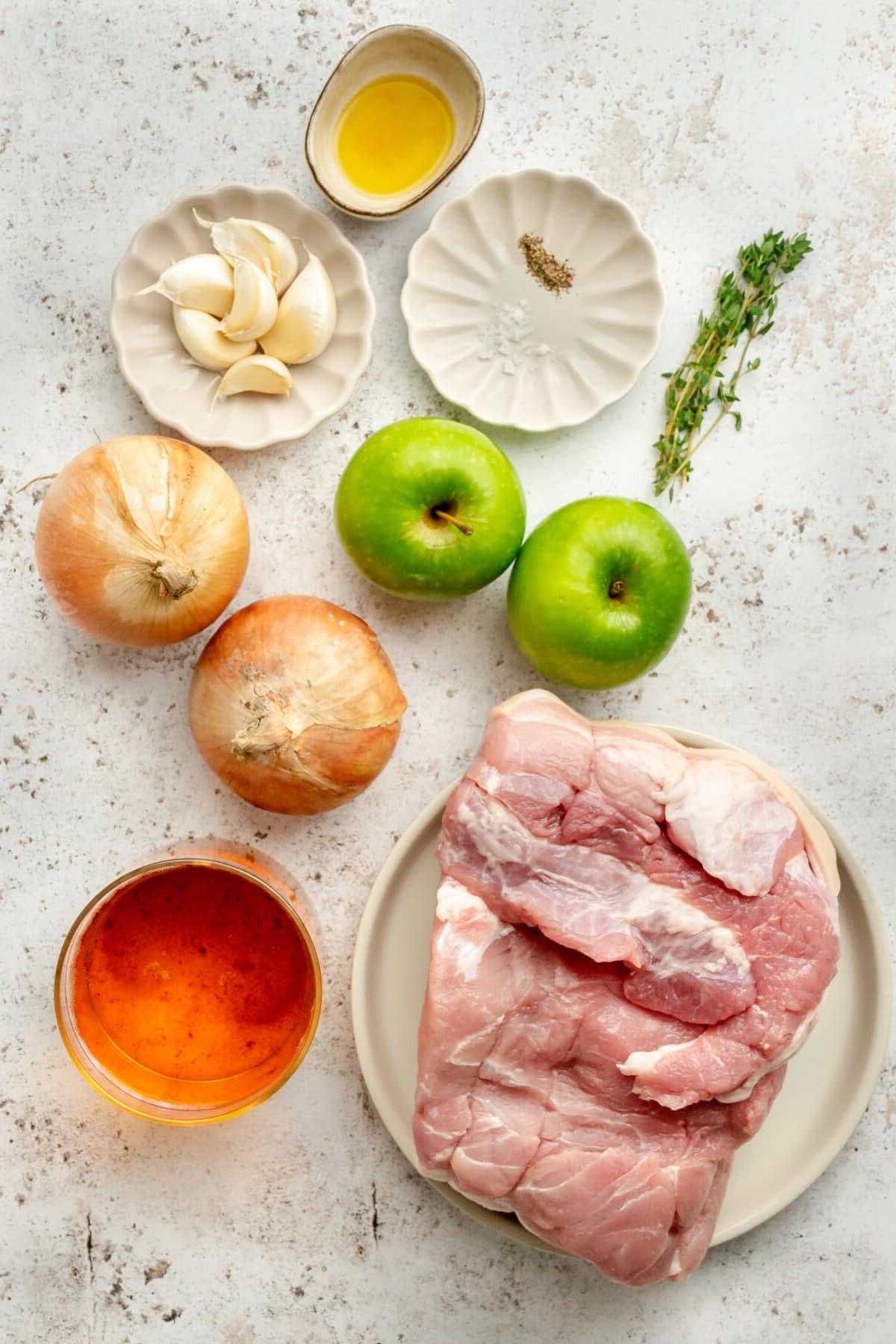 Zutaten für Schweinefiletbraten mit Apfelwein, in verschiedenen Schüsseln und auf Tellern auf einer hellgrauen Oberfläche arrangiert.