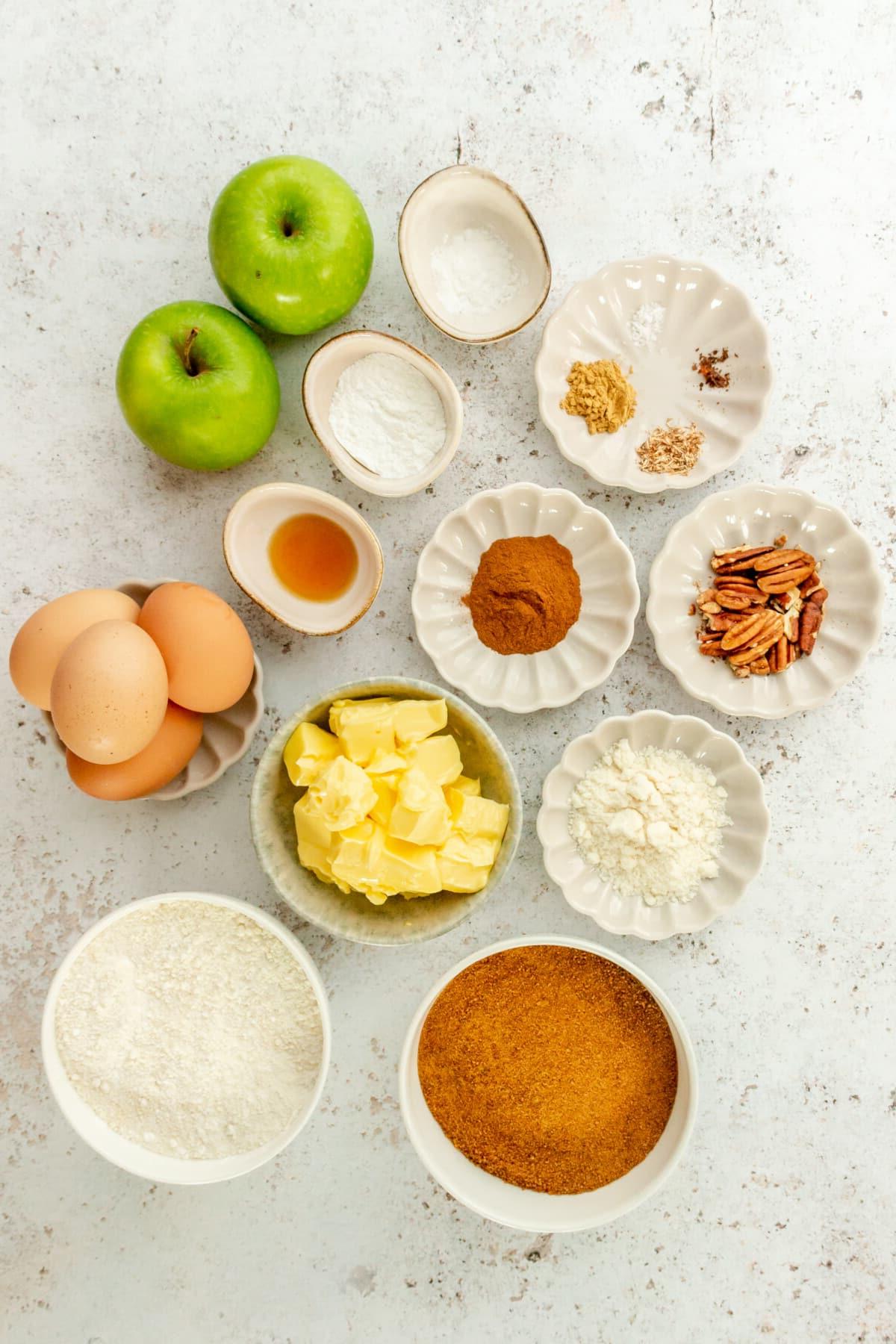 Ingredients for paleo apple cinnamon muffins sit in a variety of bowls and plates on a light grey surface.