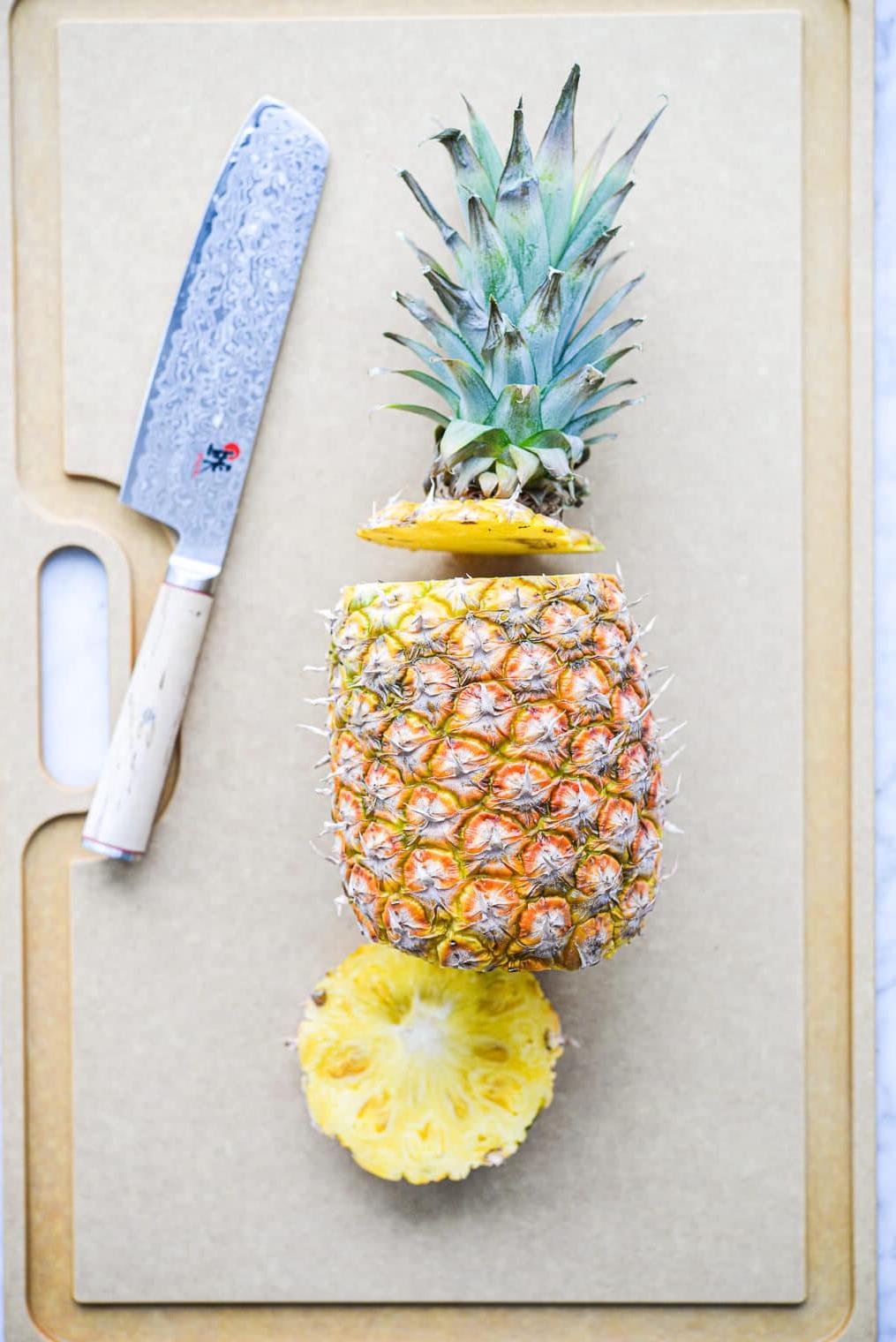 Eine Ananas liegt seitlich auf einem großen Schneidebrett, wobei der Boden und die Oberseite der Ananas abgeschnitten sind.