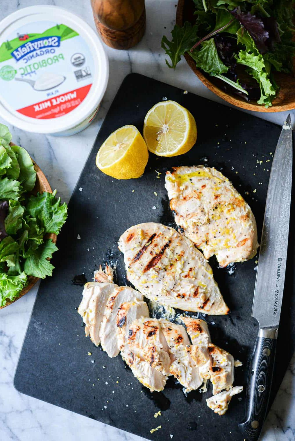 Drei Zitronen-Pfeffer-Joghurt-Hähnchenbrüste vom Grill auf einem schwarzen Schneidebrett neben einem Becher Naturjoghurt und einer halbierten Zitrone