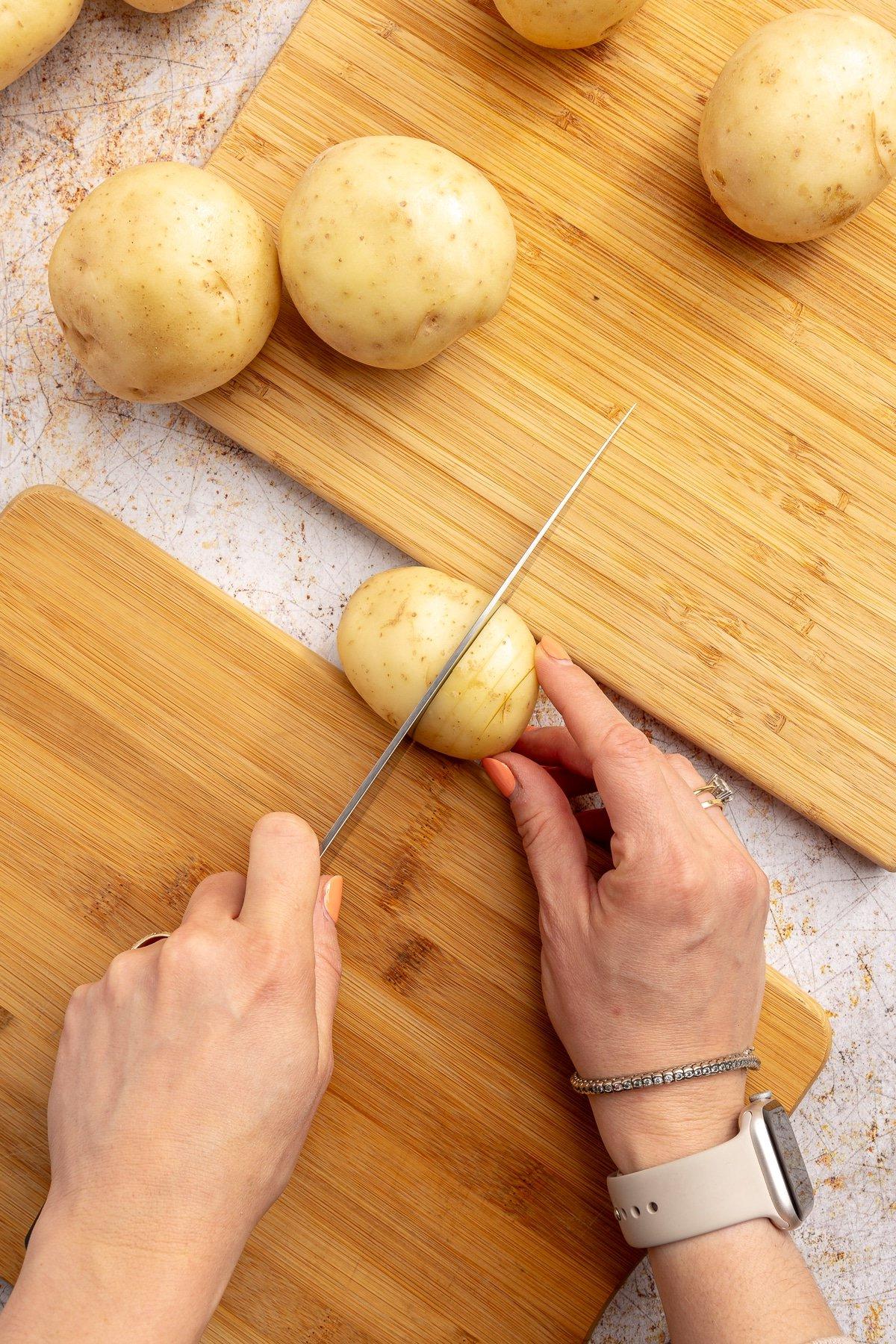 Zwei Hände schneiden eine Kartoffel, die zwischen zwei Schneidebrettern liegt. Die restlichen Kartoffeln liegen daneben.