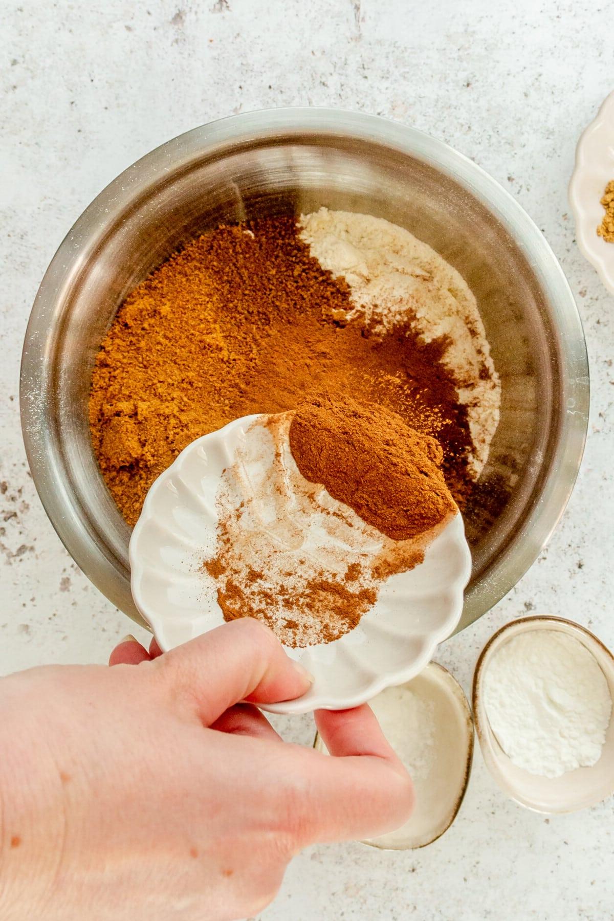 Ground cinnamon is tossed into a stainless steel bowl on a light grey surface.