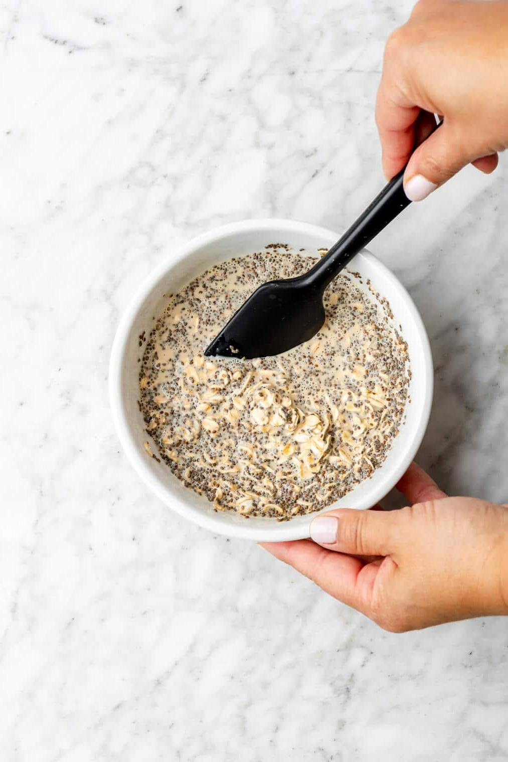 Hand rührt mit einem kleinen schwarzen Spatel Haferflocken, Chia und Milch in einer Schüssel um.