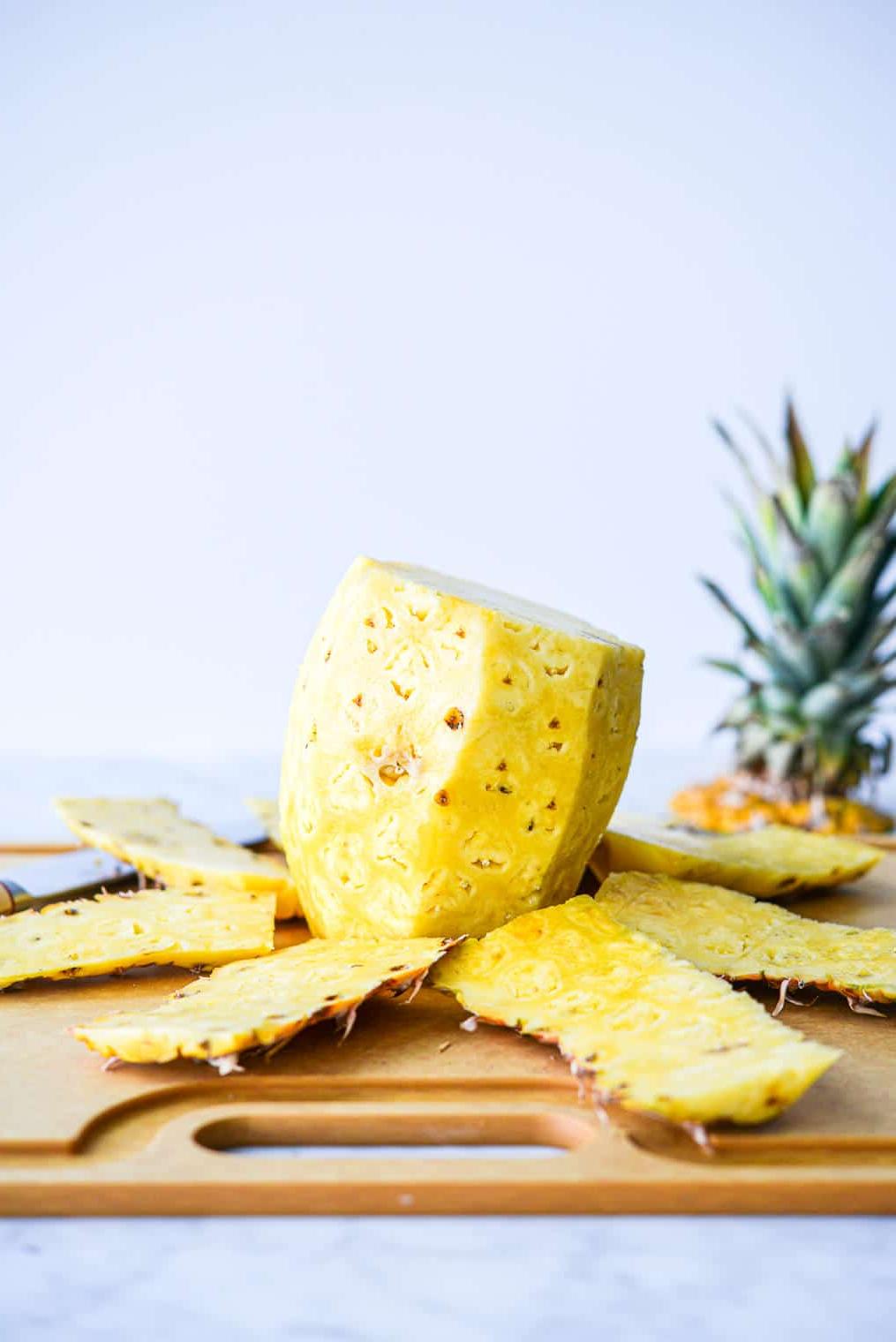 Eine Ananas, deren Schale entfernt wurde, liegt neben dem Kern auf einem Schneidebrett neben einem großen Messer.