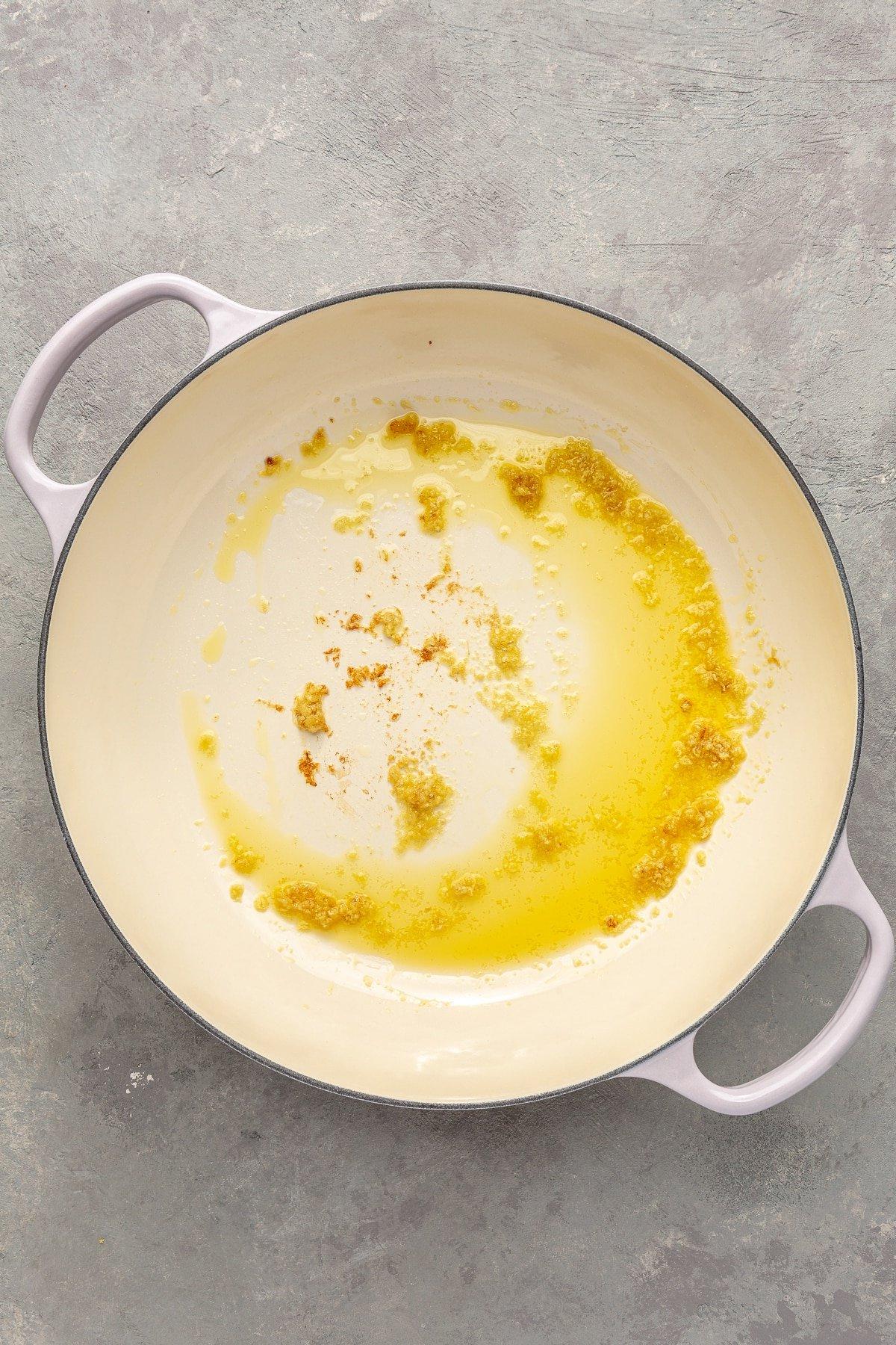 Einfaches Kürbiscurry-Rezept 4 Ghee und gehackter Knoblauch wurden in einen weißen emaillierten Topf gegeben. Das Ghee ist geschmolzen und der gehackte Knoblauch leicht goldbraun geröstet.