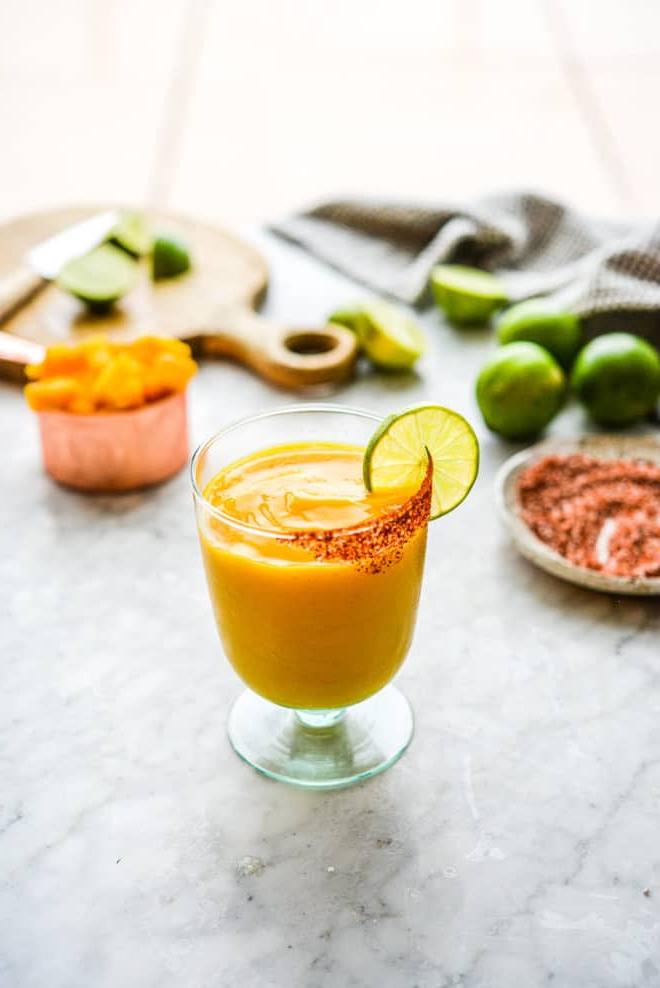 Ein Glas Frozen Mango Margarita mit Chilischnee-Rand, Zutaten für Mango Margaritas im Hintergrund