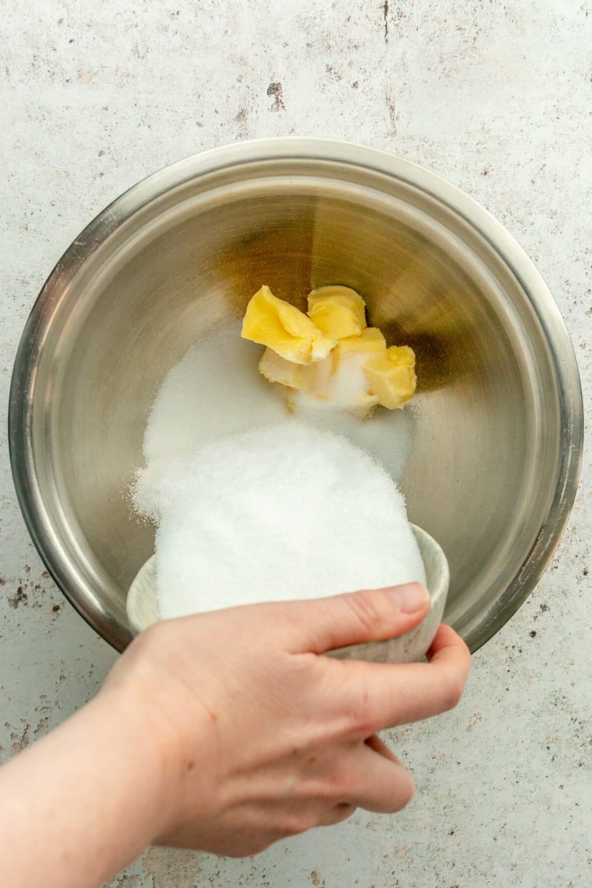 Zucker wird über Butterwürfel in einer Edelstahlschüssel auf hellem grauem Untergrund gegeben.