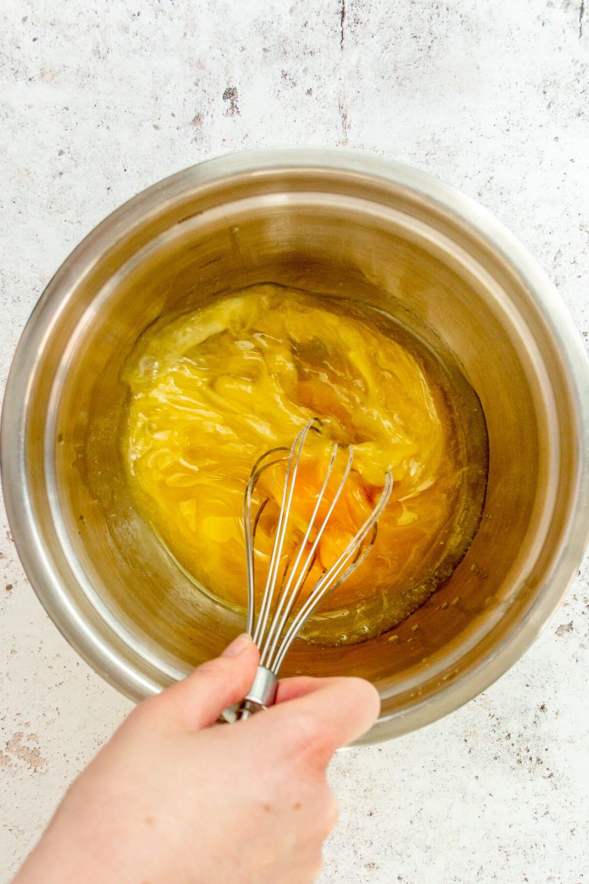 An egg and butter mixture is whisked in a stainless steel bowl on a light grey surface.