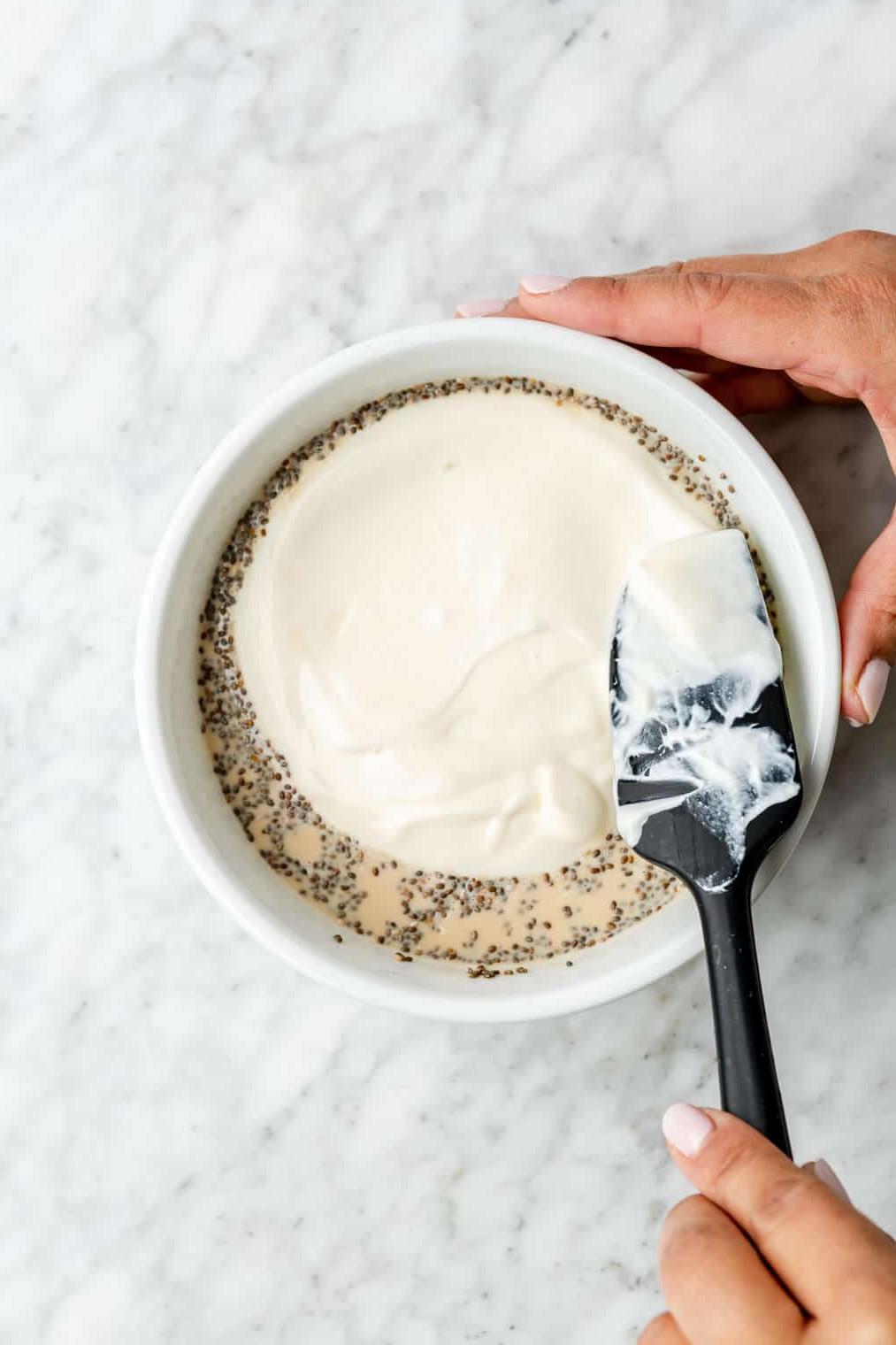 Hand streicht mit einem schwarzen Spatel Joghurt über die Haferflockenmischung in einer Schüssel.