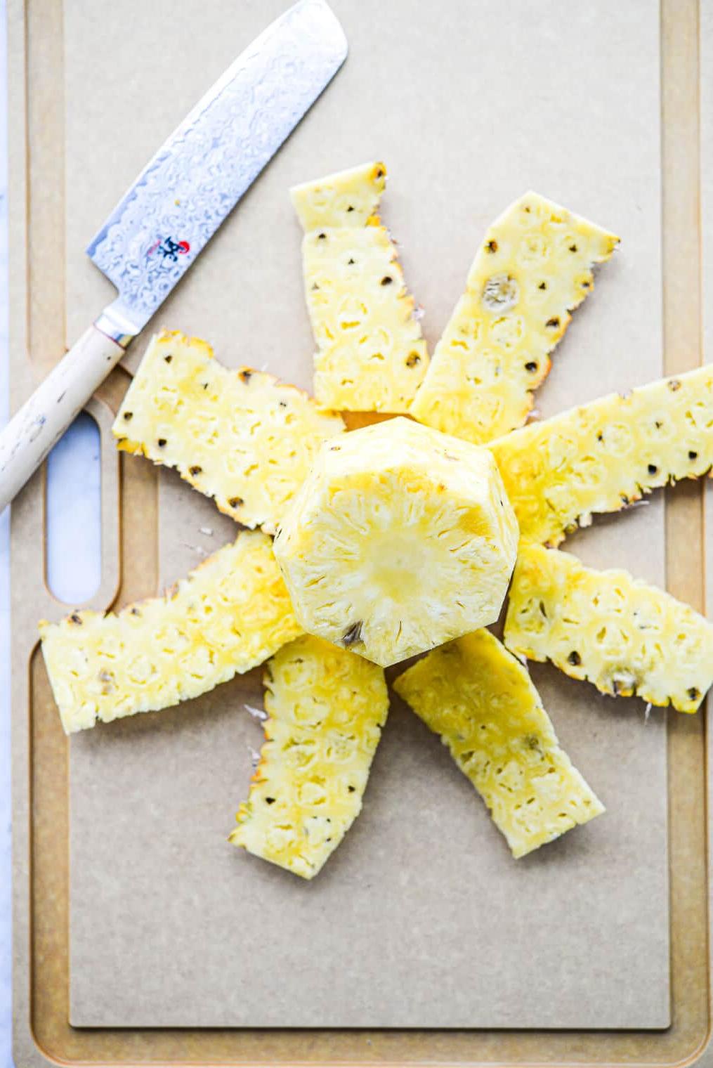 Eine Ananas, deren Schale entfernt wurde, liegt neben dem Kern auf einem Schneidebrett neben einem großen Messer.