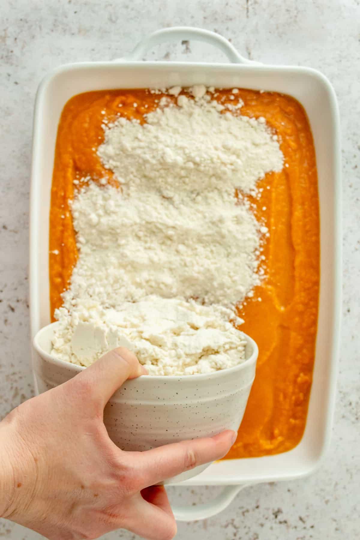 Eine gelbe Kuchenmischung wird über eine Kürbiskuchenmischung in einer weißen, rechteckigen Auflaufform auf hellgrauem Untergrund gestreut.