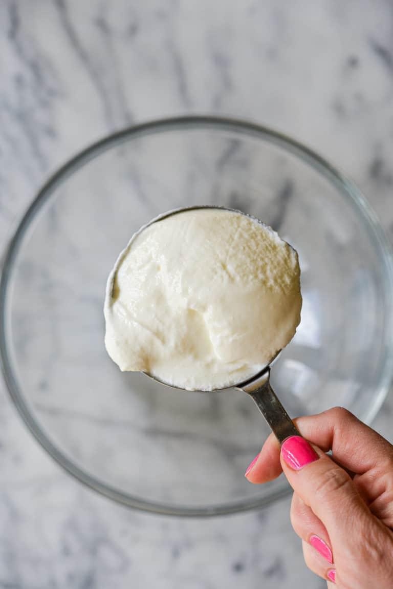 Ein Messbecher voll Naturjoghurt über einer leeren, durchsichtigen Schüssel auf einer Marmoroberfläche