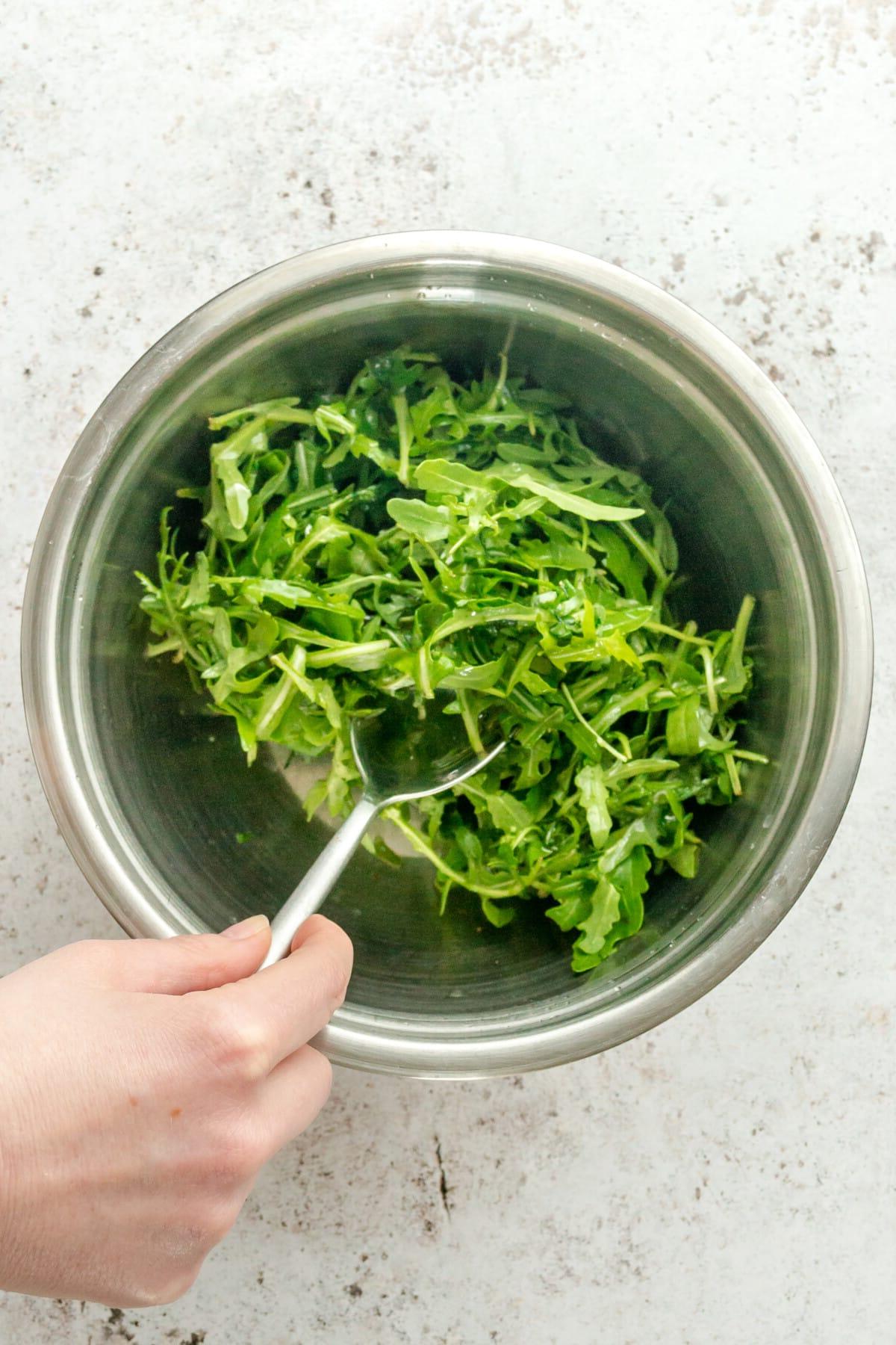 Rucola Salat mit Zitrone und Meersalz 5 Rucola wird mit einem leichten Dressing in einer Edelstahlschüssel auf hellem Hintergrund vermischt.