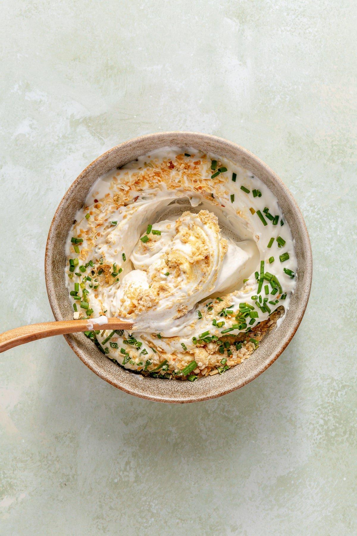 Eine Schüssel mit griechischem Joghurt, Sauerrahm und allen Gewürzen, die für hausgemachten Französischen Zwiebeldip benötigt werden, und eine Person, die die Zutaten mit einem Löffel zusammenrührt.