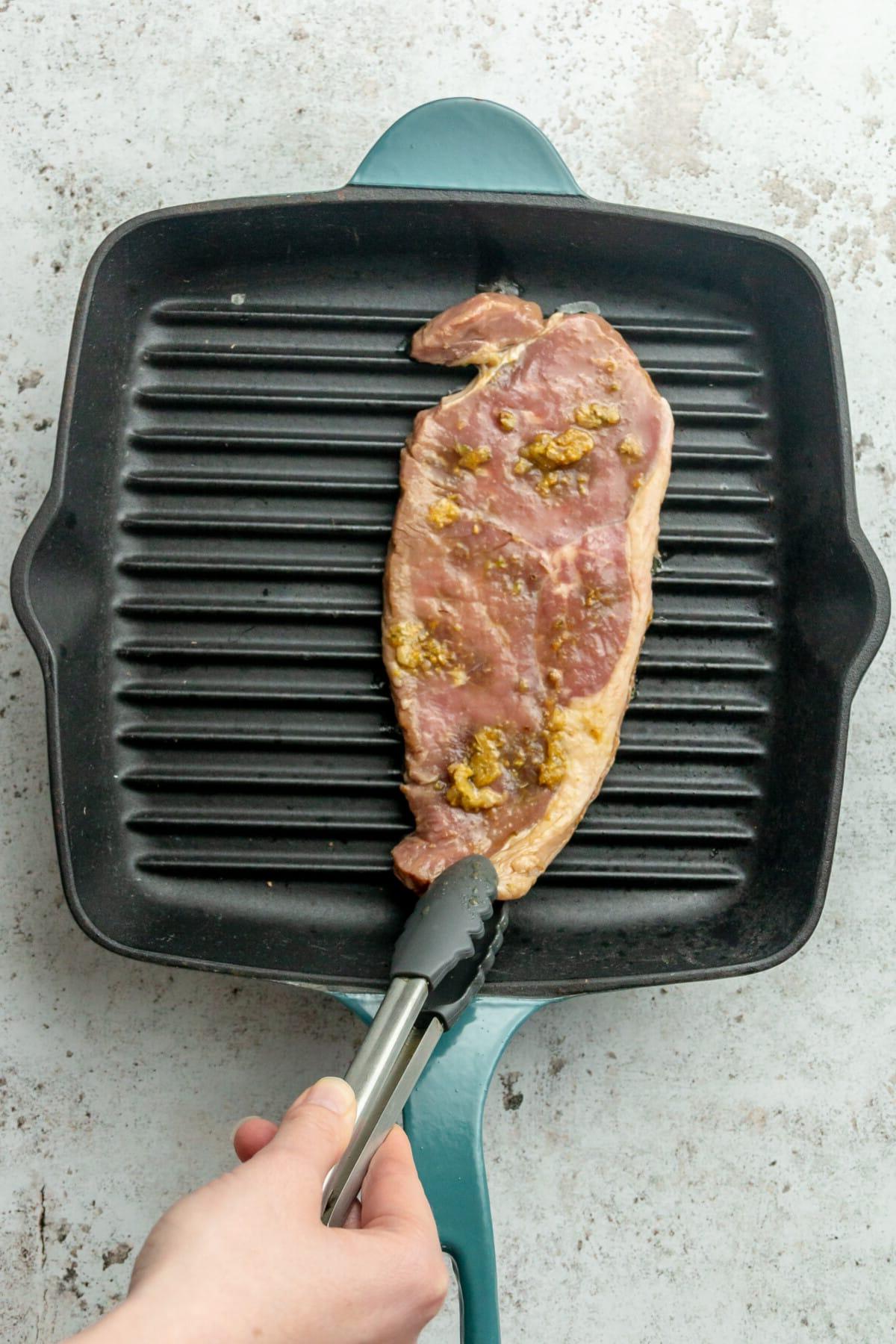 Ein Stück Steak wird auf eine heiße Grillpfanne auf einer hellgrauen Oberfläche gelegt.