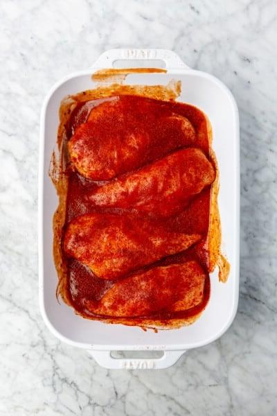 4 rohe Hähnchenbrüste, bedeckt mit BBQ-Sauce und Gewürzen in einer weißen Auflaufform.