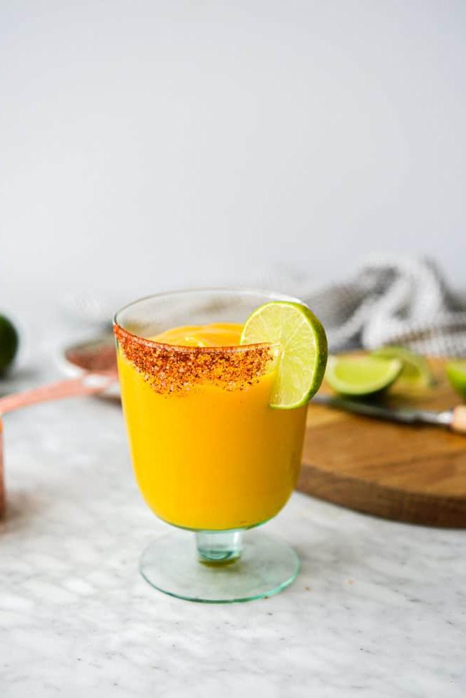 Ein Glas Frozen Mango Margarita mit Chilischnee-Rand, Zutaten für Mango Margaritas im Hintergrund
