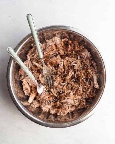 210414_Carnitas-30 Eine Schüssel mit zerkleinertem Schweinefleisch, bestreut mit Gewürzen, und zwei Gabeln in der Schüssel zum Mischen