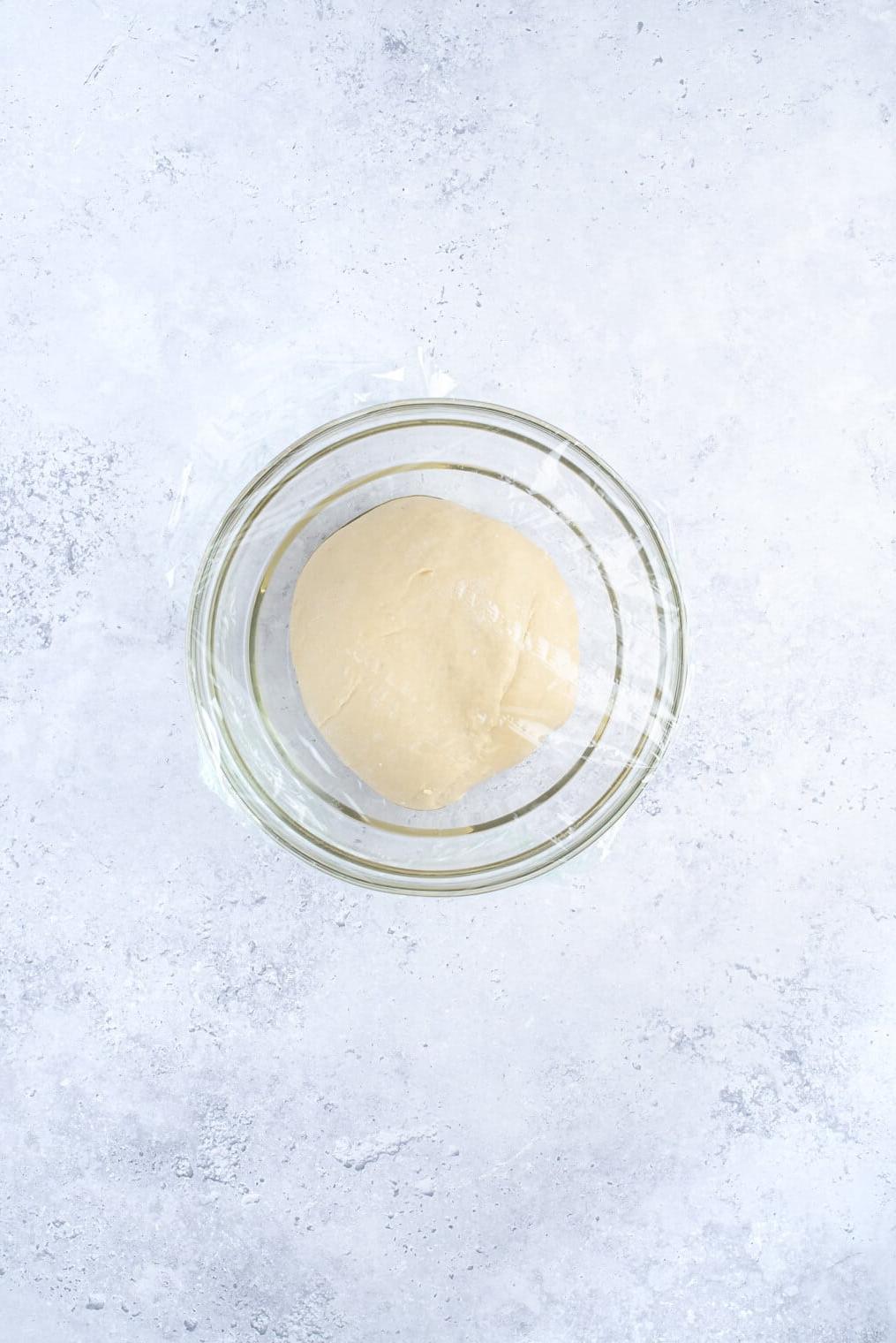 Veganer Zimtschnecken-Teig geht in einer mit Frischhaltefolie abgedeckten Glasschale auf