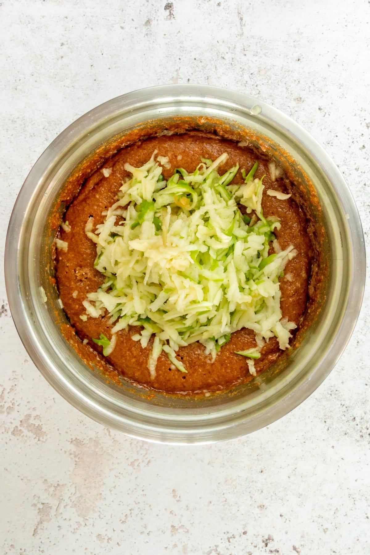 Grated apple sits on top of a paleo cinnamon muffin batter in a stainless steel bowl on a light grey surface.